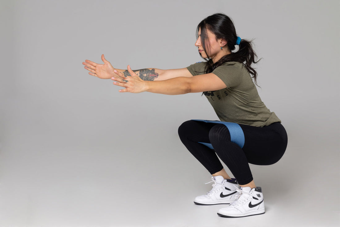 A woman has her arms extended as she lowers down into a bodyweight squat with a band wrapped around her thighs.