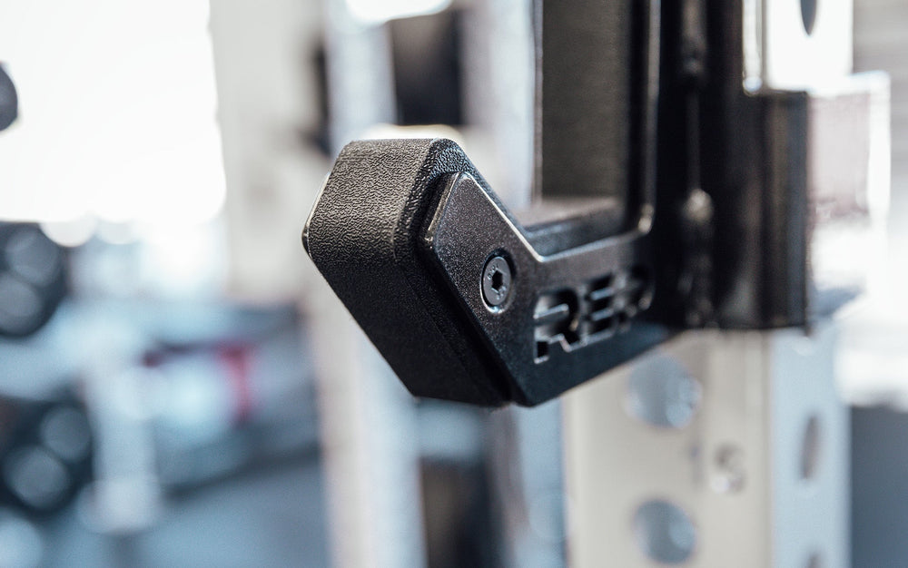 Close-up of Flat Sandwich J-Cups 2.0 – heavy-duty 1,000 lb capacity barbell holders for PR-5000 power racks mounted on white rack upright; 3x3” steel design with urethane and plastic protection, laser-cut REP logo plate, stainless-steel backing, metallic black powder coat, and wider opening for specialty bars from Rep Fitness UK | Shop Premium Gym Equipment for Home & Commercial Use, Best Online Gym Equipment UK