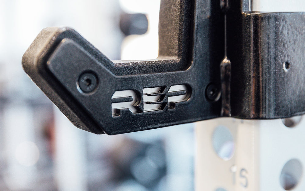 Close-up of REP logo on Flat Sandwich J-Cups 2.0 – heavy-duty 1,000 lb capacity barbell holders for PR-5000 power racks mounted on white rack upright; 3x3” steel design with urethane and plastic protection, laser-cut REP logo plate, stainless-steel backing, metallic black powder coat, and wider opening for specialty bars from Rep Fitness UK | Shop Premium Gym Equipment for Home & Commercial Use, Best Online Gym Equipment UK