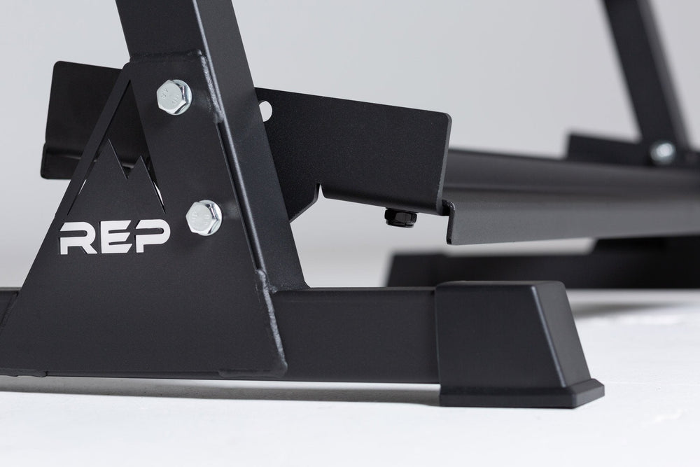 Close-up side view of the bottom of the 3-tiered matte black REP Dumbbell Rack showcasing the "REP" logo, bottom shelf, and rubber foot. 3-tiered matte black REP Dumbbell Rack storing a 5-50 REP dumbbell set, heavy-duty three-tier dumbbell rack with durable 11-gauge steel shelves, compact footprint, and 1" lip for secure storage  fits REP rubber hex and urethane dumbbells, available in multiple colors with easy zinc-coated hardware assembly from Rep Fitness UK | Shop Premium Gym Equipment UK online