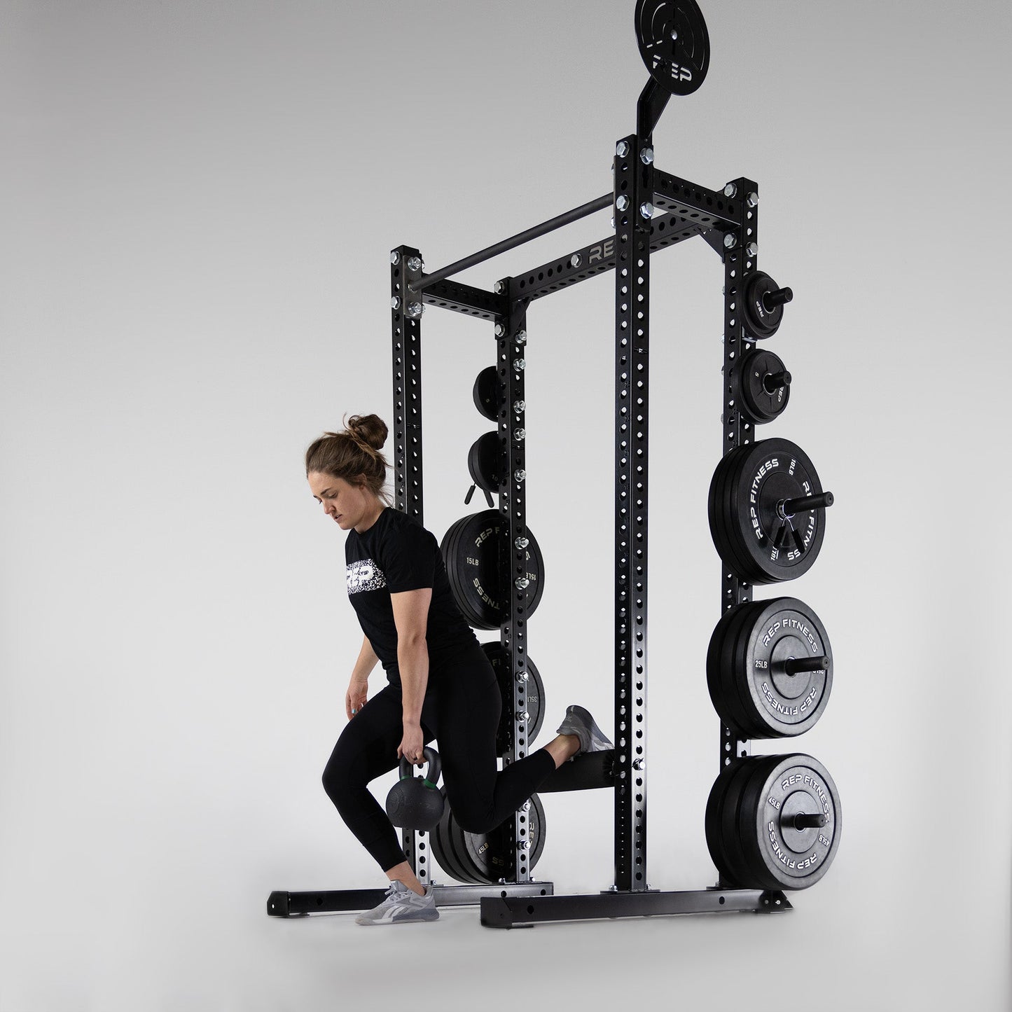 Compact Logo Plate Crossmember Shown on Power Rack being used by woman, Compact Logo Plate Crossmember Shown on power rack, heavy-duty 3x3” 11-gauge steel rack stabilizer with laser-cut holes, metallic black powder coat, and laser-engraved REP logo from Rep Fitness UK | Shop Premium Gym Equipment for Home & Commercial Use, Best Online Gym UK Online