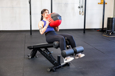Athlete doing sit ups with the Adjustable Bench Leg Roller Attachment 2.0 and a medicine ball from Rep Fitness UK | Shop Best Premium Gym Equipment for Home & Commercial Use Online
