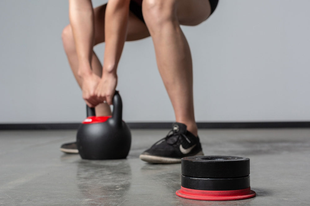 Exercising with the adjustable kettlebell with detachable weight in the foreground from Rep Fitness UK, quick weight adjustment, non-slip matte powder coat, rubber base, and versatile for home gyms| Shop Premium Gym Equipment for Home & Commercial Use, Best Online Gym Equipment UK