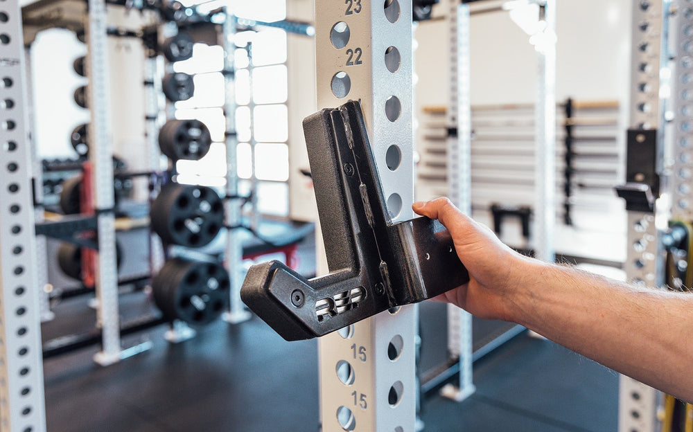 Flat Sandwich J-Cups 2.0 – heavy-duty 1,000 lb capacity barbell holders for PR-5000 power racks mounted on white rack upright; 3x3” steel design with urethane and plastic protection, laser-cut REP logo plate, stainless-steel backing, metallic black powder coat, and wider opening for specialty bars from Rep Fitness UK | Shop Premium Gym Equipment for Home & Commercial Use, Best Online Gym Equipment UK