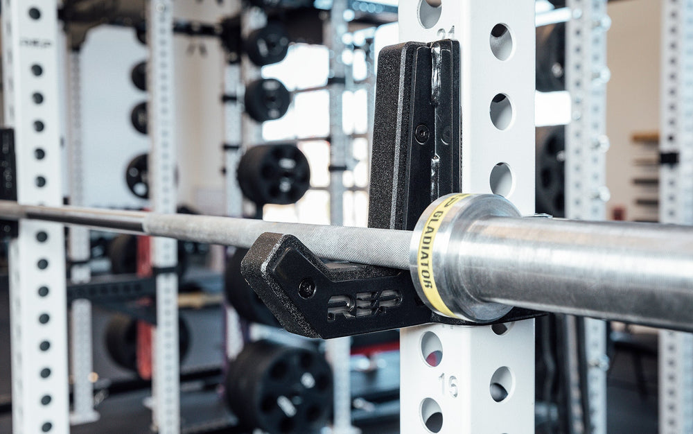 Flat Sandwich J-Cups 2.0 – heavy-duty 1,000 lb capacity barbell holders for PR-5000 power racks mounted on white rack upright with bar in place; 3x3” steel design with urethane and plastic protection, laser-cut REP logo plate, stainless-steel backing, metallic black powder coat, and wider opening for specialty bars from Rep Fitness UK | Shop Premium Gym Equipment for Home & Commercial Use, Best Online Gym Equipment UK