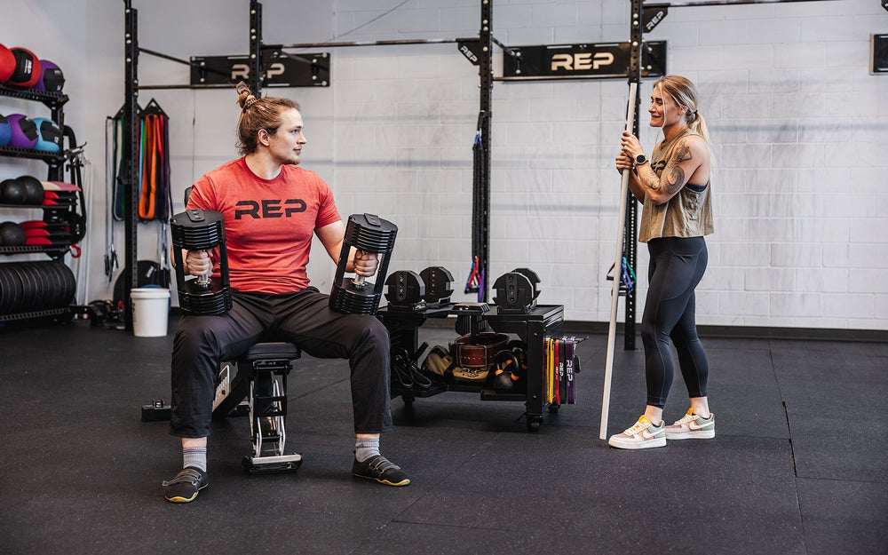 Man and woman in gym next to premium heavy duty matte black storage cart with wheels in sleek matte black metal finish for organized gym equipment storage loaded with exercise balls, kettlebells and other lifting and exercise equipment from Rep Fitness UK | Shop Premium Gym Equipment for Home & Commercial Use Online
