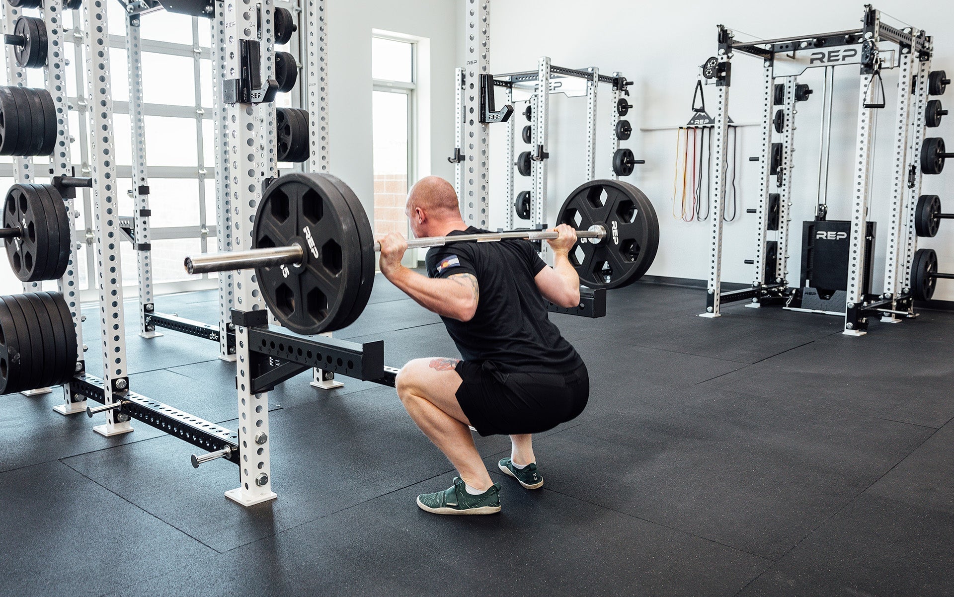 Man doing squats with REP Fully Stainless Steel Double Black Diamond Power Bar, elite 20kg powerlifting barbell with ultra-aggressive mountain knurling, 29mm stiff shaft, bronze bushings, and IPF-standard specs, built for maximum grip and durability; available in stainless steel or Cerakote finishes for premium performance and corrosion resistance from Rep Fitness UK | Shop Premium Gym Equipment for Home & Commercial Use, Best UK Gym Equipment Online