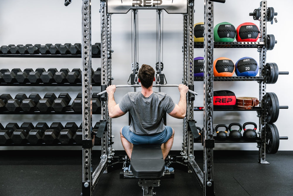 Man exercising surrounded by Ball & Plate Storage Shelfs, heavy-duty 11-gauge steel gym storage shelf for bumper plates, medicine balls, and slam balls, 600lb capacity, 41” wide with laser-cut REP logo, metallic black powder-coat finish, compatible with PR-5000 power rack for organized, space-saving gym setup from Rep Fitness UK | Shop Premium Gym Equipment for Home & Commercial Use, Best Online Gym Equipment UK