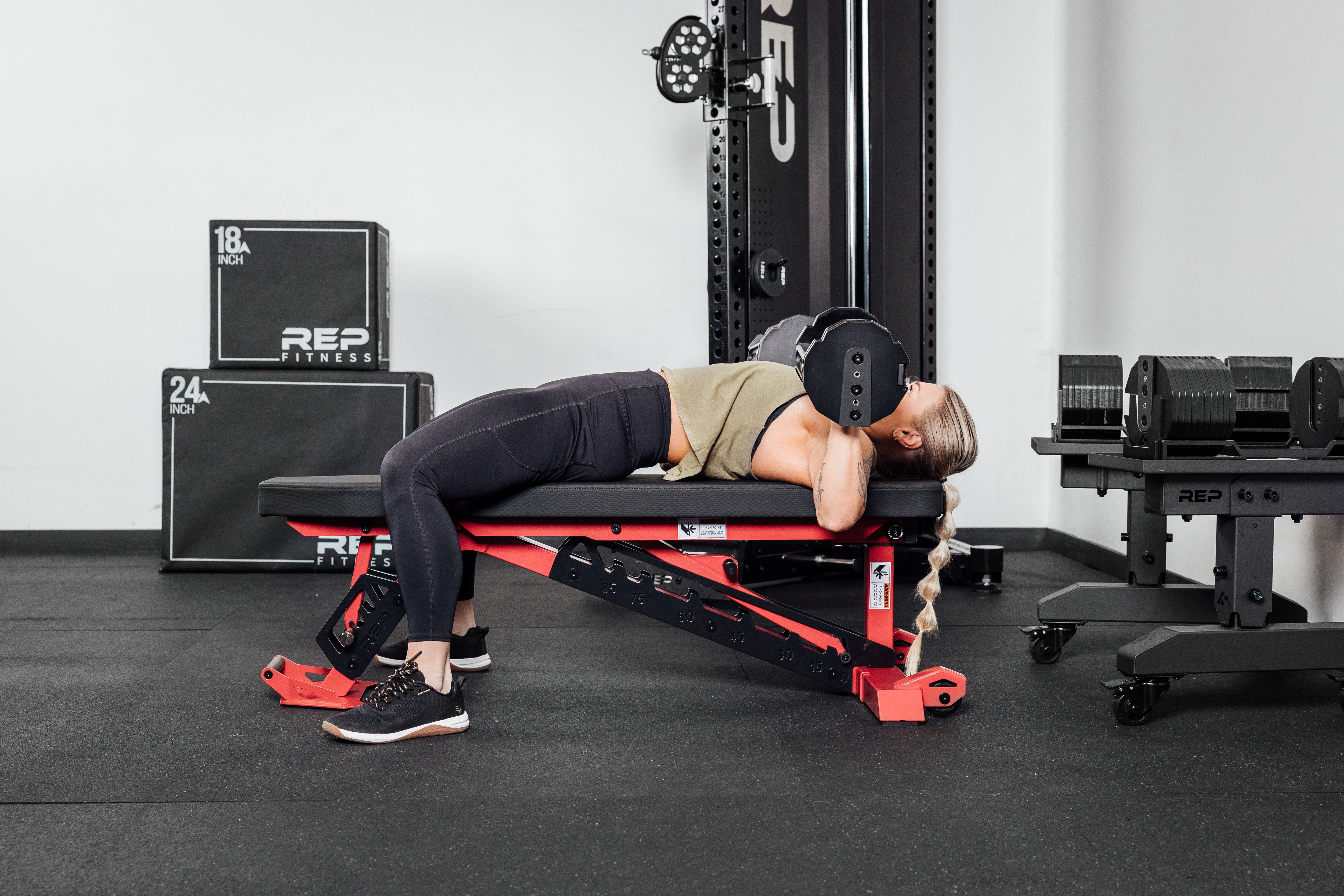 Nighthawk exercise bench red woman using Pepin dumbbells from Rep Fitness UK | Shop Premium Gym Equipment for Home & Commercial Use Online compact, high-performance bench for home and light-commercial gyms; 7- and 14-gauge steel frame, 7 back and 4 seat positions, CleanGrip™ vinyl pad, knurled handle, heavy-duty wheels, IPF-compliant 16.7” height, vertical storage, and compatibility with leg curl and roller attachments from Rep Fitness UK | Shop Best Premium Gym Equipment for Home & Commercial Use Online