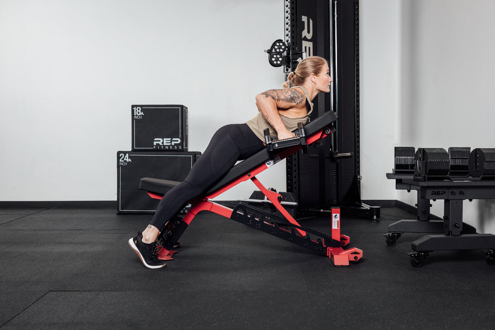 Nighthawk exercise bench red woman laying on using Pepin dumbbells from Rep Fitness UK | Shop Premium Gym Equipment for Home & Commercial Use Online compact, high-performance bench for home and light-commercial gyms; 7- and 14-gauge steel frame, 7 back and 4 seat positions, CleanGrip™ vinyl pad, knurled handle, heavy-duty wheels, IPF-compliant 16.7” height, vertical storage, and compatibility with leg curl and roller attachments from Rep Fitness UK | Shop Best Premium Gym Equipment for Home & Commercial Use