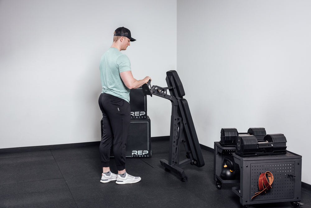 Nighthawk matte black bench man rolling folded bench into corner for tidy storage from Rep Fitness UK | Shop Premium Gym Equipment for Home & Commercial Use Online