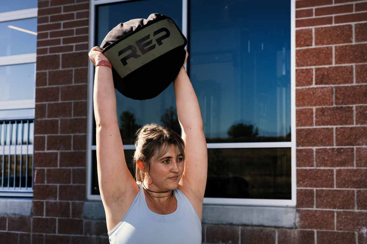 Person lifting a weighted  stone sandbag with 'REP' branding outdoors overhead