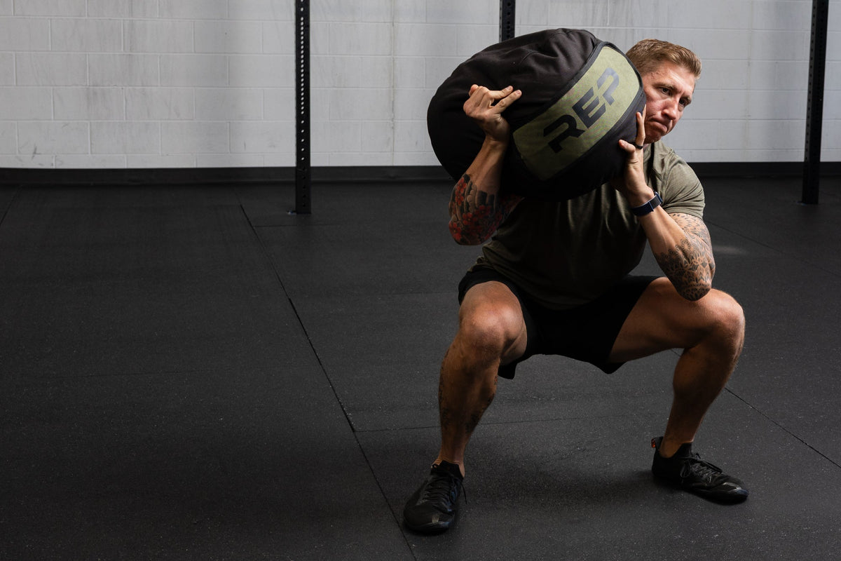 Person performing a squat with a weighted stone sandbag in a gym setting