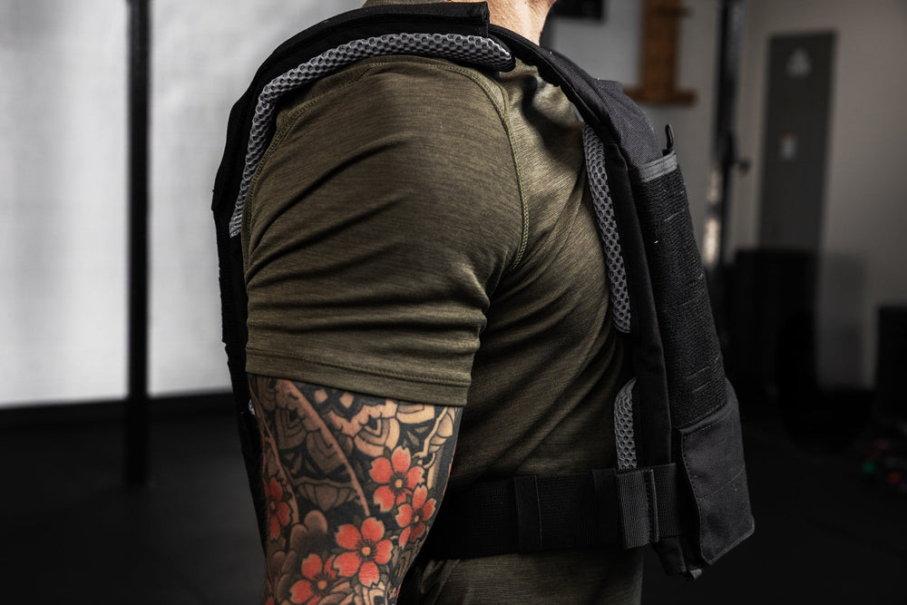 Person wearing a green shirt and black vest with a tattoo on their arm, standing indoors showing the thickness of the vest with the soft weight vest plate