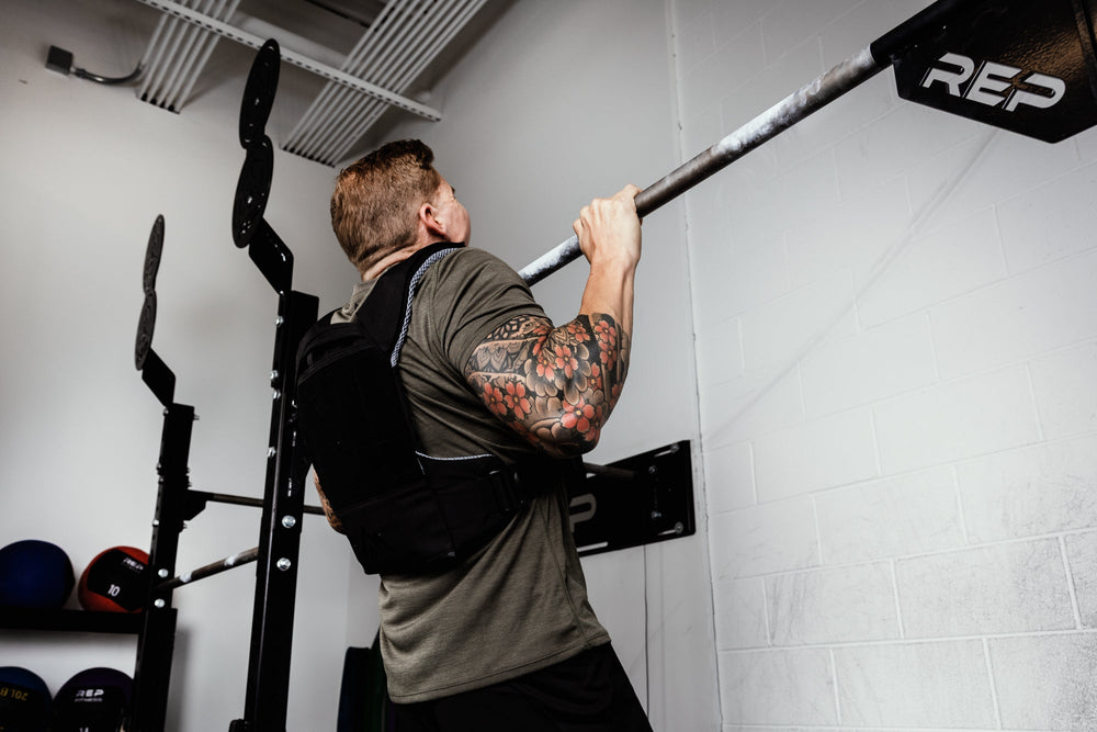 Person performing pull-ups on a bar with 'REP' branding  weight vest and soft weight vest plate in a gym setting.