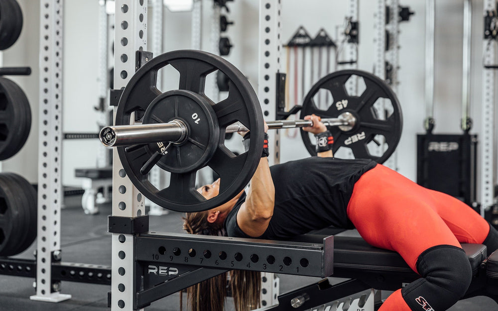 Woman doing bench press using REP Fully Stainless Steel Double Black Diamond Power Bar, elite 20kg powerlifting barbell with ultra-aggressive mountain knurling, 29mm stiff shaft, bronze bushings, and IPF-standard specs, built for maximum grip and durability; available in stainless steel or Cerakote finishes for premium performance and corrosion resistance from Rep Fitness UK | Shop Premium Gym Equipment for Home & Commercial Use, Best UK Gym Equipment Online