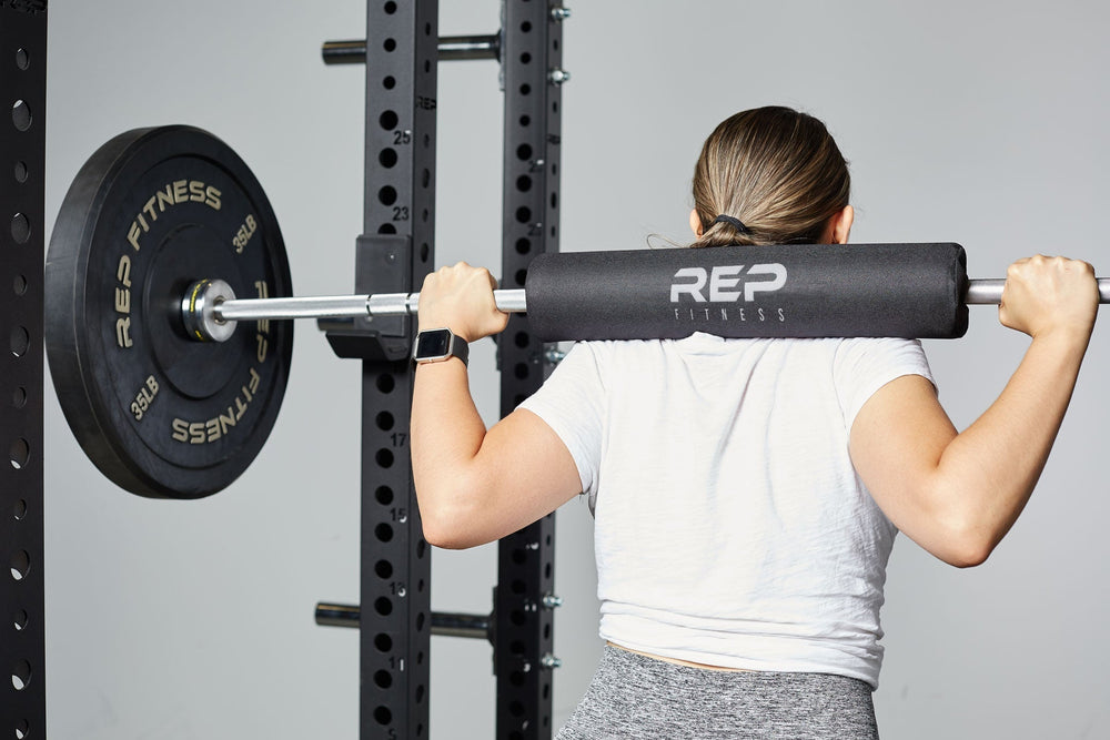Woman holding barbell with Barbell Shoulder Pad on shoulders, soft, durable squat and hip thrust pad with Velcro closure for easy attachment; protects shoulders, neck, and hips during lifts; made from nitrile rubber and Oxford cloth; 17” long, 4” diameter with white REP Fitness logo stitching for comfort and stylefrom Rep Fitness UK | Shop Premium Gym Equipment for Home & Commercial Use, Best Online Gym Equipment UK