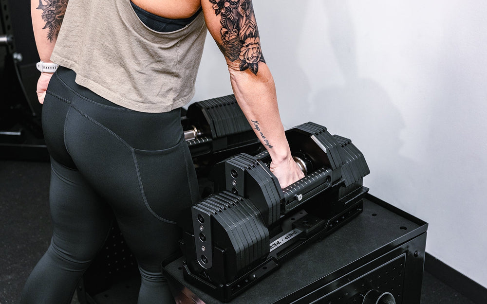 Woman lifting Pepin dumbbell from compact multipurpose storage cart for weights and accessories in matte black metal finish for organized gym equipment storage from Rep Fitness UK | Shop Premium Gym Equipment for Home & Commercial Use Online
