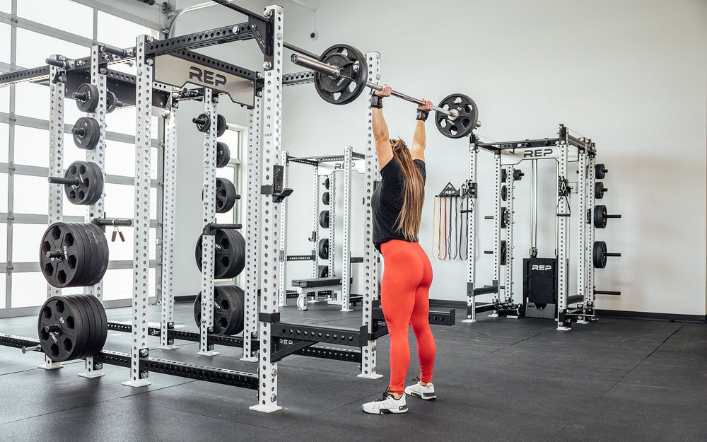 Woman lifting above head using REP Fully Stainless Steel Double Black Diamond Power Bar, elite 20kg powerlifting barbell with ultra-aggressive mountain knurling, 29mm stiff shaft, bronze bushings, and IPF-standard specs, built for maximum grip and durability; available in stainless steel or Cerakote finishes for premium performance and corrosion resistance from Rep Fitness UK | Shop Premium Gym Equipment for Home & Commercial Use, Best UK Gym Equipment Online