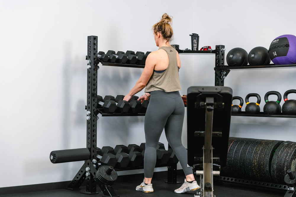 Woman lifting up two dumbbells from a Dumbbell Storage Shelf – heavy-duty 11-gauge steel dumbbell rack shelf with 600lb capacity, metallic black powder-coat finish, laser-cut REP logo, compatible with 4000 and 5000 Series racks, fits full 5–50lb dumbbell sets for organized, space-saving gym storage from Rep Fitness UK | Shop Premium Gym Equipment for Home & Commercial Use, Best Online Gym Equipment UK