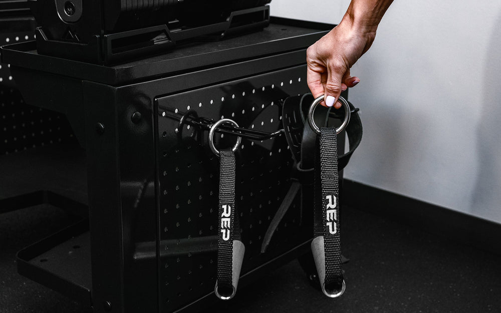 Woman picking up pulley handle hanging from heavy-duty multipurpose cart  storage solution in sleek matte black metal finish for organized gym equipment storage from Rep Fitness UK | Shop Premium Gym Equipment for Home & Commercial Use Online