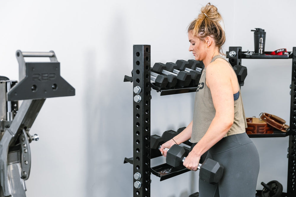 Woman picking up two dumbbells from a Dumbbell Storage Shelf – heavy-duty 11-gauge steel dumbbell rack shelf with 600lb capacity, metallic black powder-coat finish, laser-cut REP logo, compatible with 4000 and 5000 Series racks, fits full 5–50lb dumbbell sets for organized, space-saving gym storage from Rep Fitness UK | Shop Premium Gym Equipment for Home & Commercial Use, Best Online Gym Equipment UK