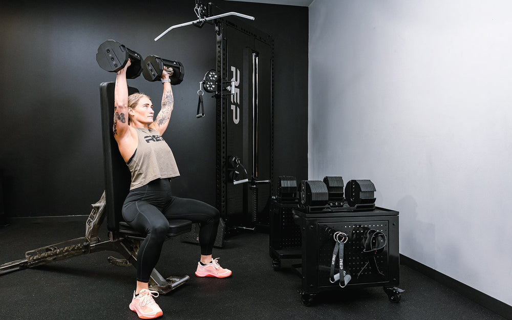 Woman sitting lifting dumbbells next to durable matte black storage cart rack with wheels in sleek matte black metal finish for organized gym equipment storage from Rep Fitness UK | Shop Premium Gym Equipment for Home & Commercial Use Online