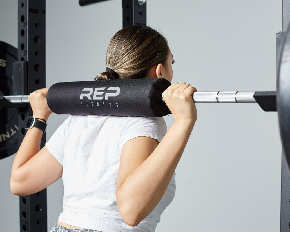 Woman with Barbell Shoulder Pad on barbell on shoulders, soft, durable squat and hip thrust pad with Velcro closure for easy attachment; protects shoulders, neck, and hips during lifts; made from nitrile rubber and Oxford cloth; 17” long, 4” diameter with white REP Fitness logo stitching for comfort and stylefrom Rep Fitness UK | Shop Premium Gym Equipment for Home & Commercial Use, Best Online Gym Equipment UK