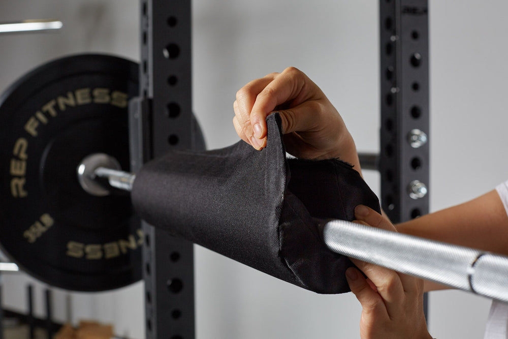 Woman wrapping Fitness Barbell Shoulder Pad on barbell, soft, durable squat and hip thrust pad with Velcro closure for easy attachment; protects shoulders, neck, and hips during lifts; made from nitrile rubber and Oxford cloth; 17” long, 4” diameter with white REP Fitness logo stitching for comfort and stylefrom Rep Fitness UK | Shop Premium Gym Equipment for Home & Commercial Use, Best Online Gym Equipment UK